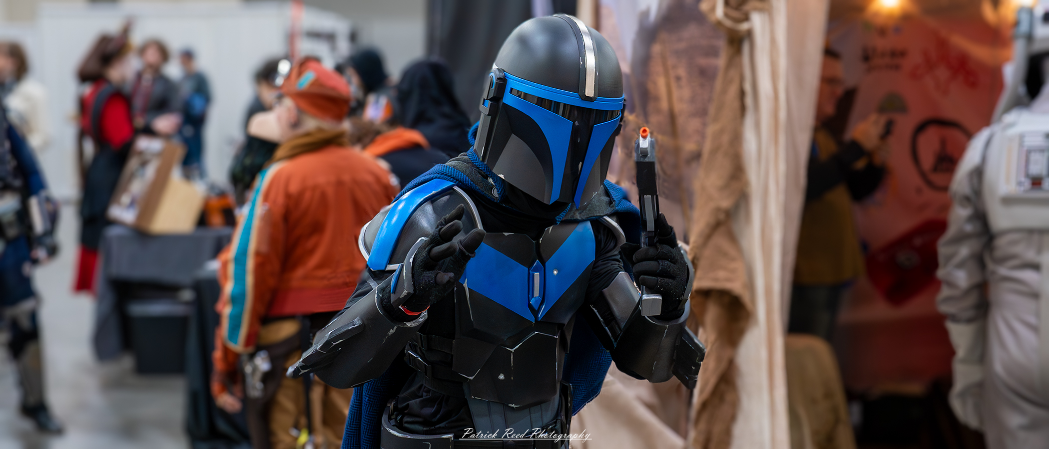A cosplayer dressed as a Mandalorian, wearing striking blue armor with a polished finish, a T-visor helmet, and a blaster holstered at their side, standing in a heroic pose that exudes the iconic bounty hunter's mystique.